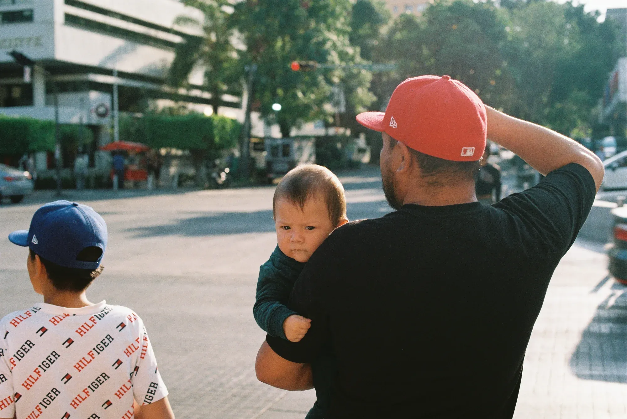 Hombre de gorra roja carga a un bebé de cara molesta en una avenida, junto a un niño de gorra azul