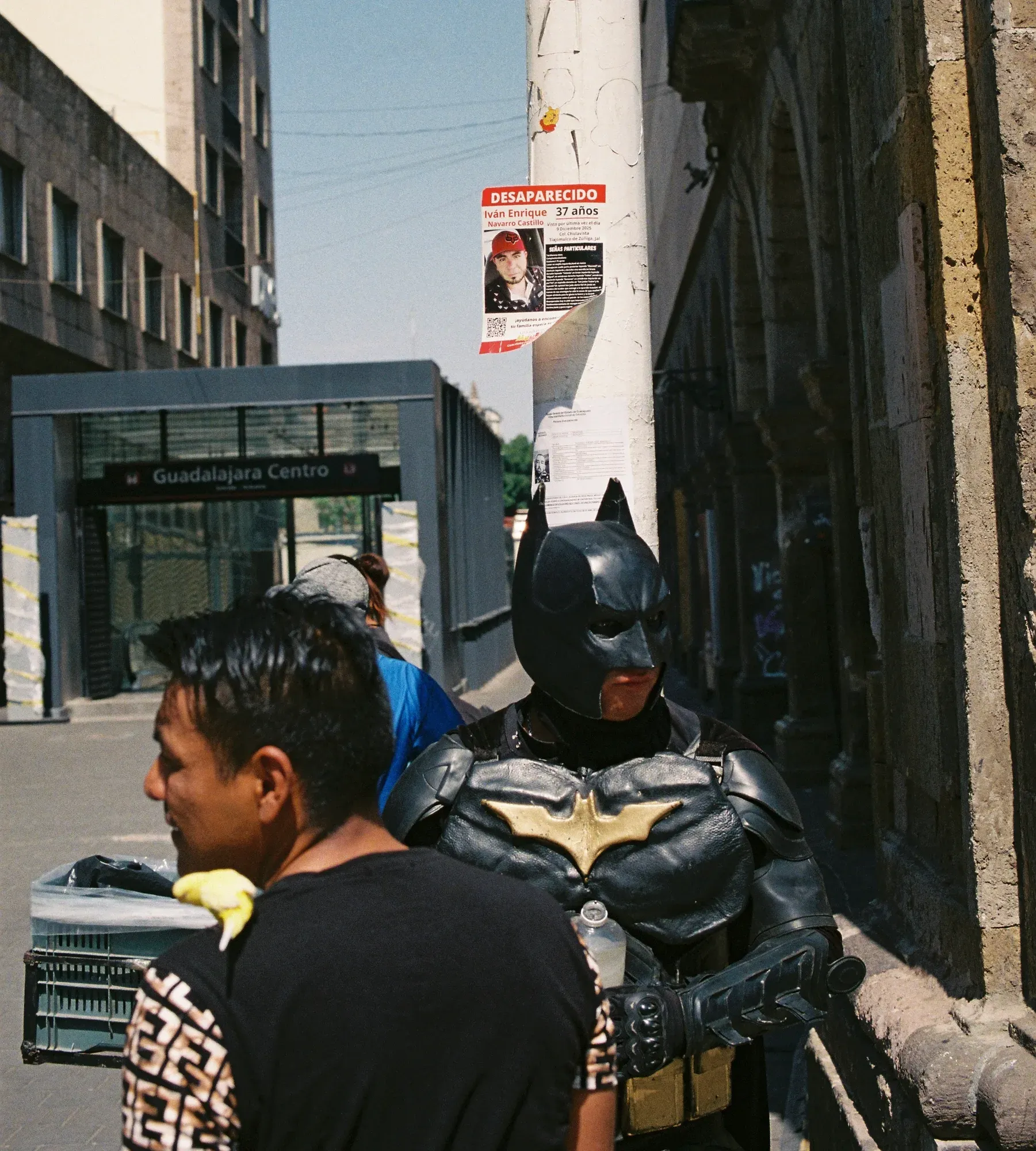 Persona disfrazada de Batman junto a un poste con un cartel de desaparecido, a la salida del metro Guadalajara Centro