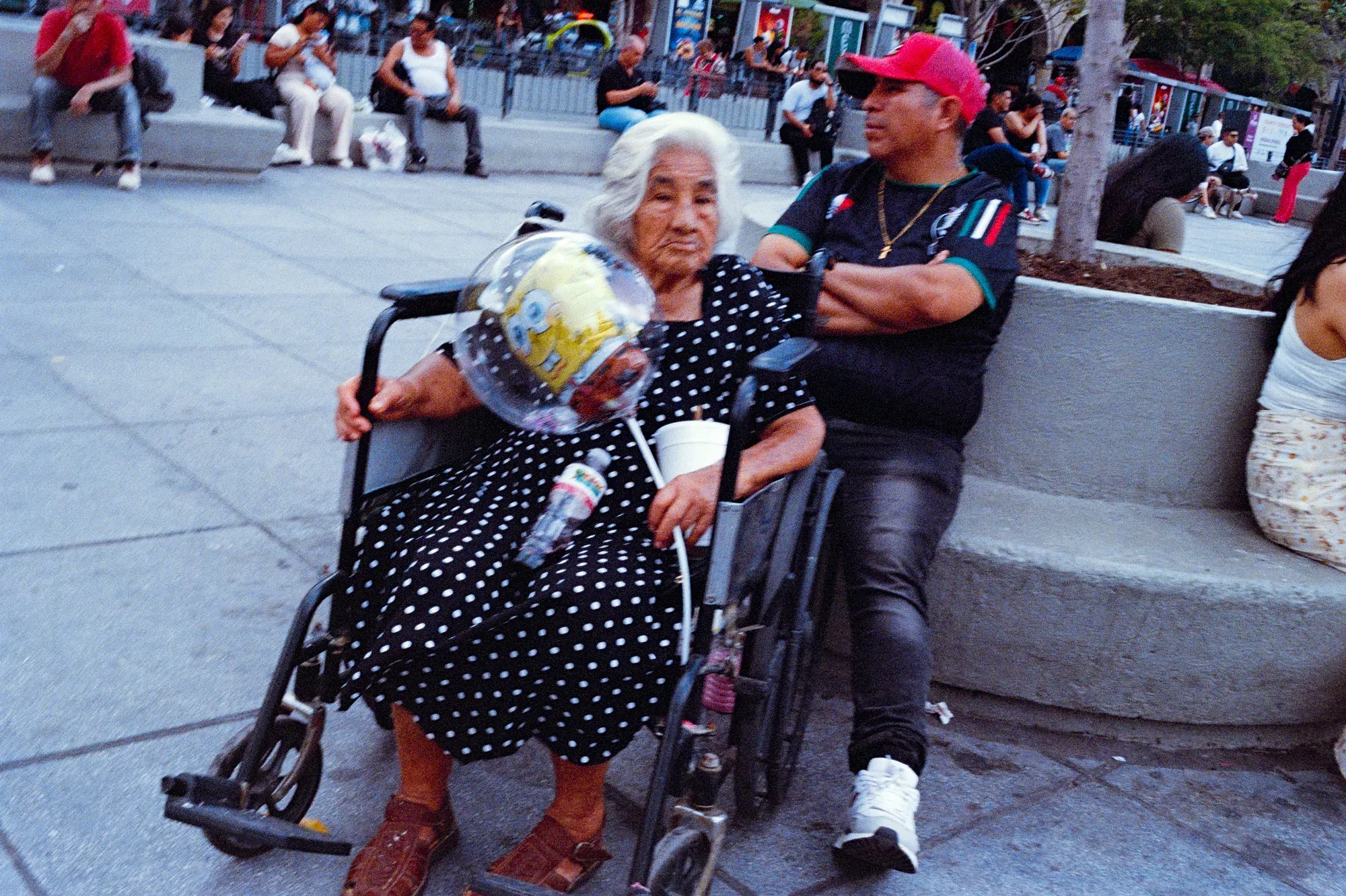 Mujer mayor en silla de ruedas con un globo en una plaza, junto a un hombre sentado en una banca
