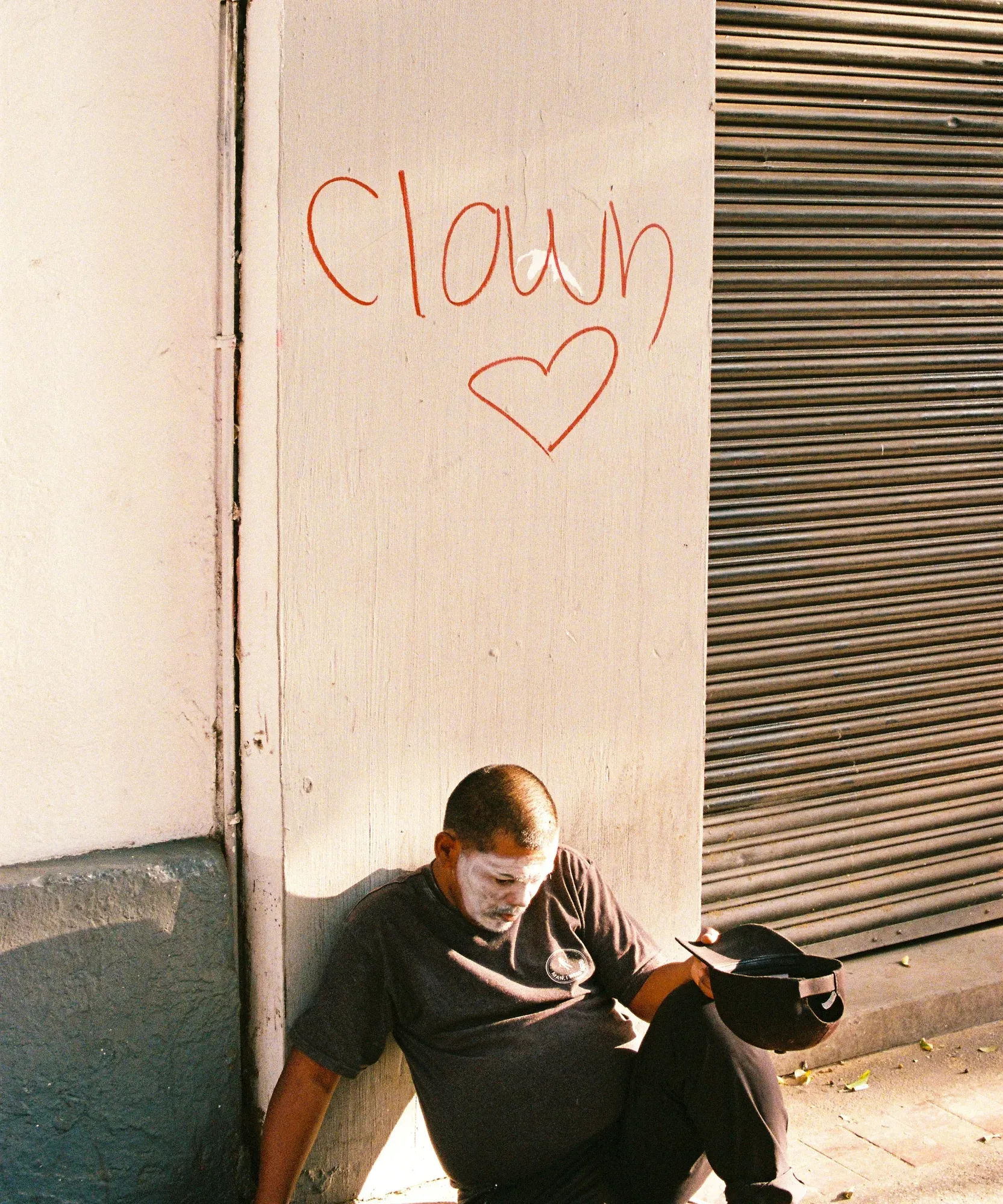 Hombre con maquillaje blanco sentado en el suelo, bajo un grafiti que dice «clown» y un corazón