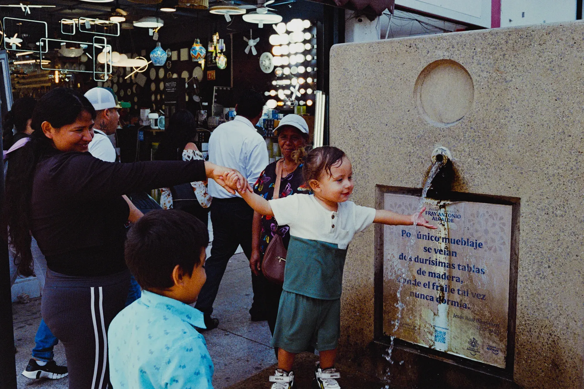 Niña juega con el chorro de una fuente de la calle, tomada de la mano de su madre