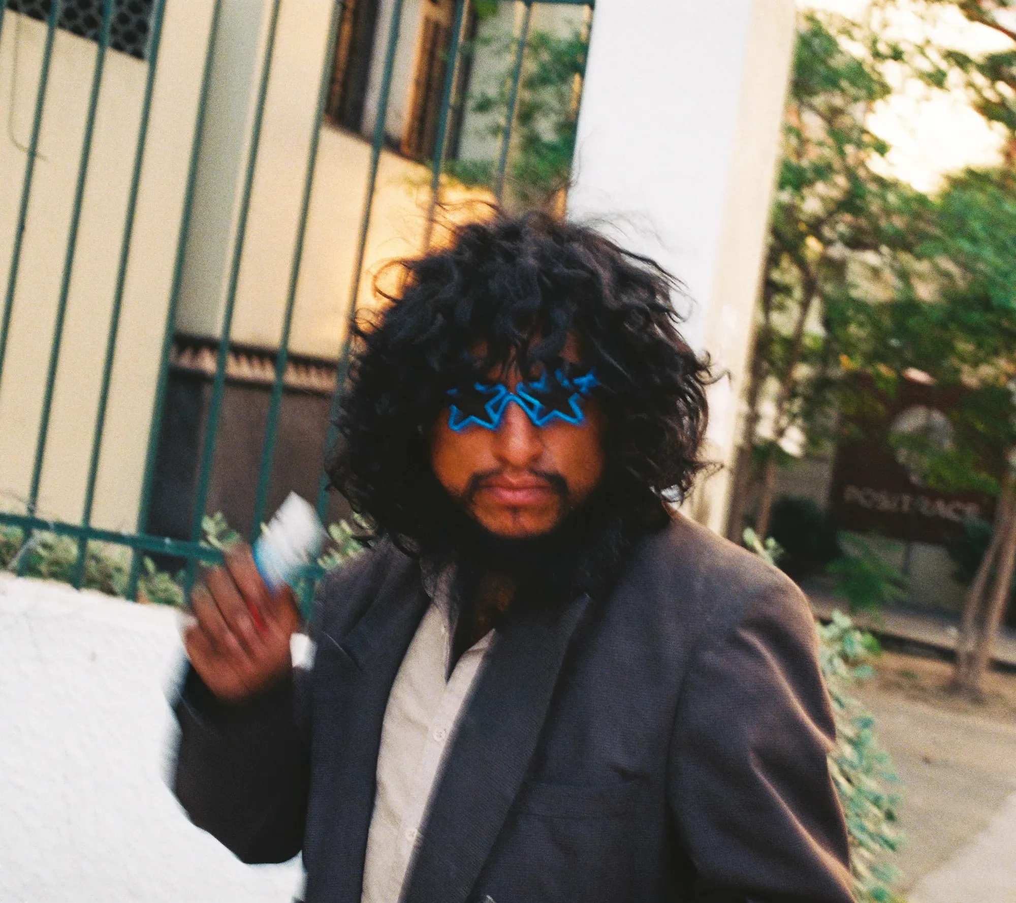 Hombre de cabello rizado con lentes de sol azules en forma de estrella, frente a una reja verde