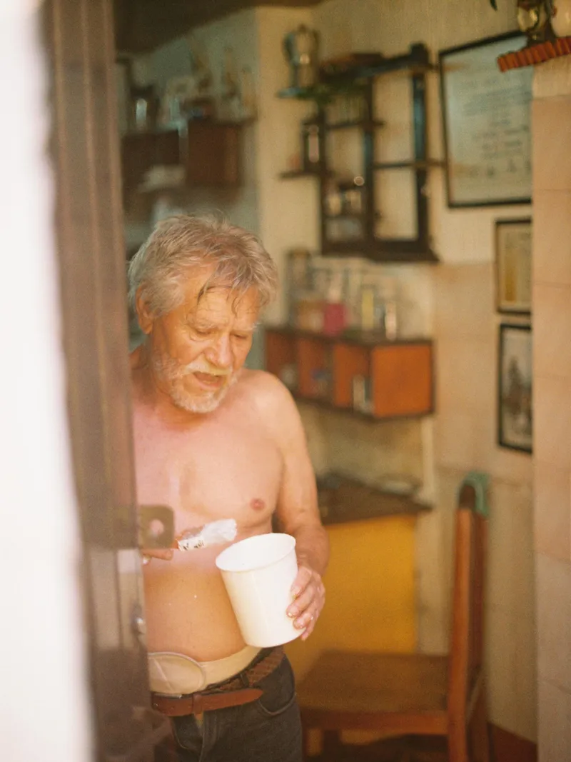 Hombre mayor sin camisa con un vaso en su cocina