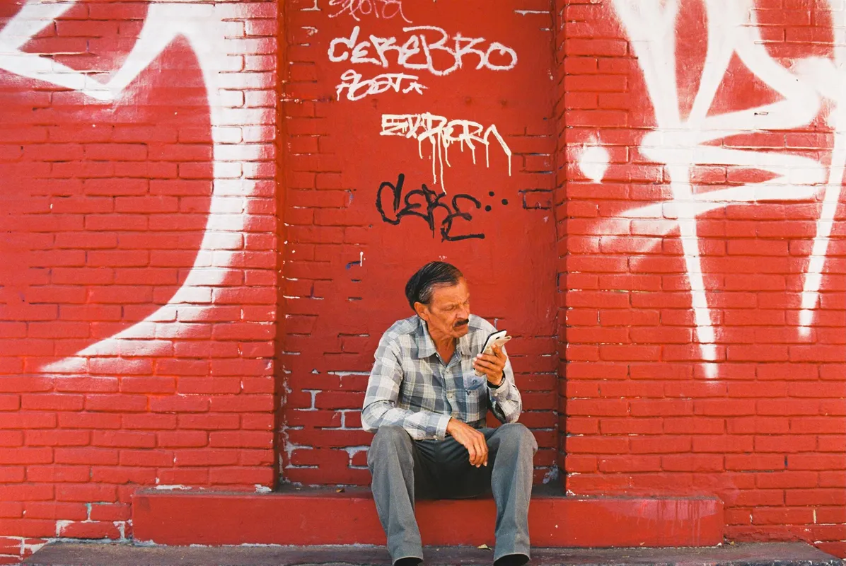 Hombre sentado frente a un muro rojo con grafiti mirando su celular