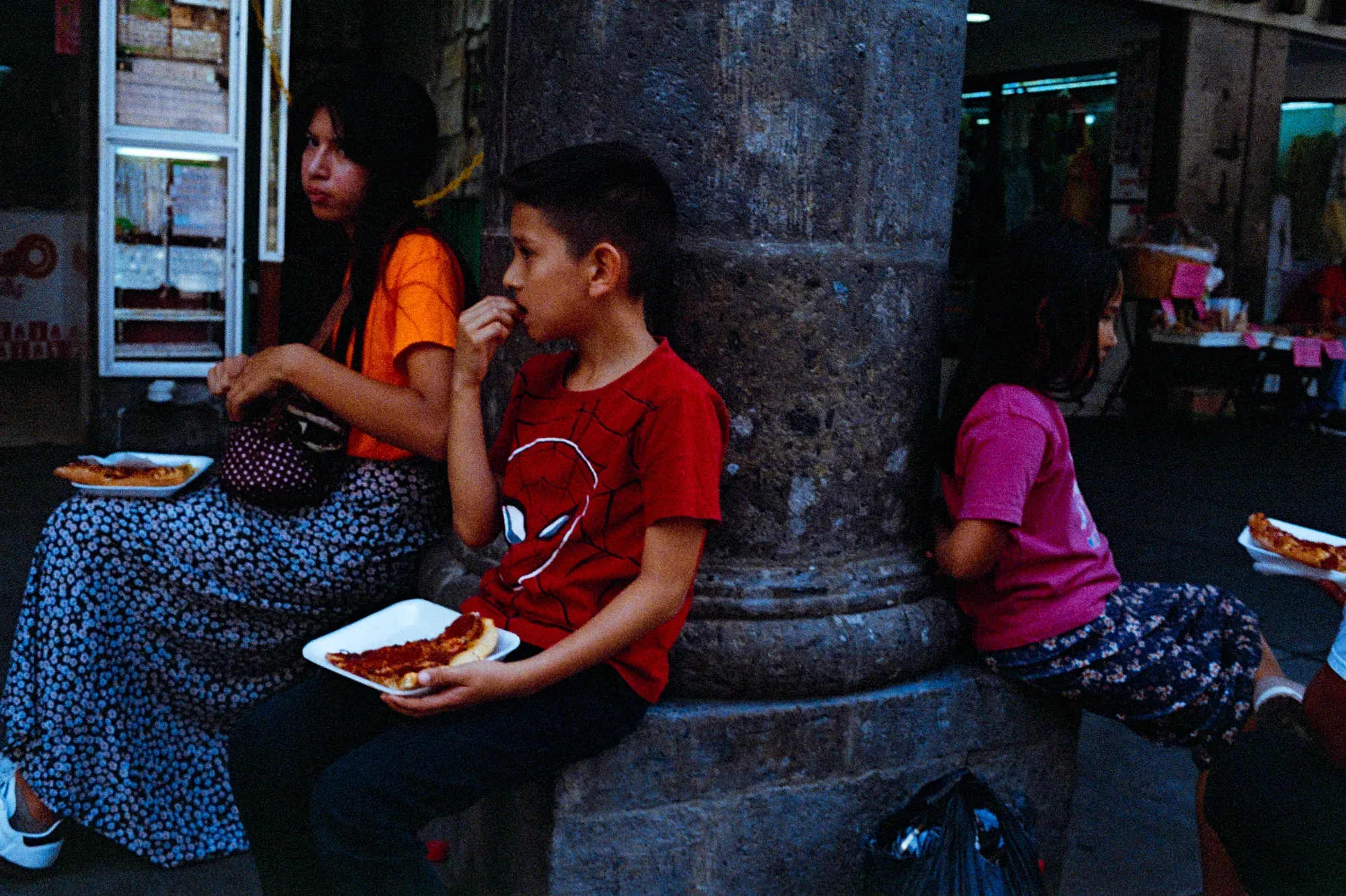 Tres personas comen pizza sentadas junto a una columna en la calle