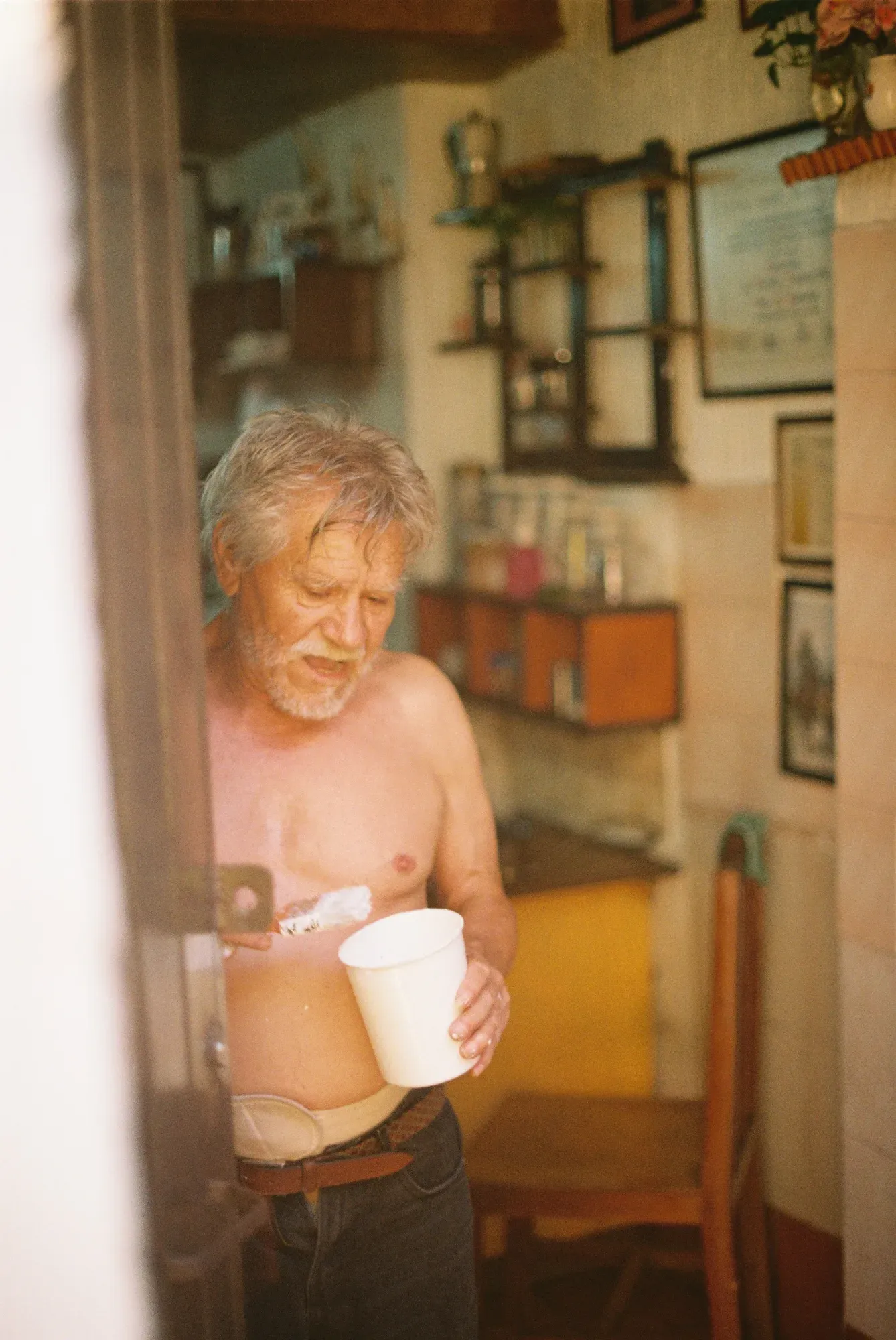 Hombre mayor sin camisa con un vaso en la mano en su cocina